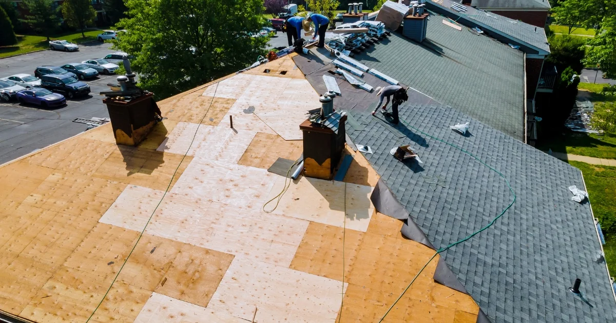 Roofers installing new shingles on a large residential roof, partially covered in plywood sheathing.